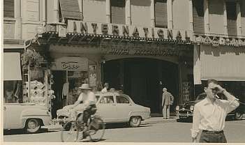 Extérieur de l'Hotel International avec des personnes, des voitures anciennes et un homme à vélo