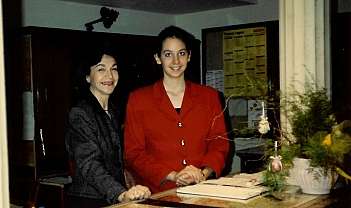 Deux femmes souriantes derrière un comptoir de réception avec des plantes et des documents