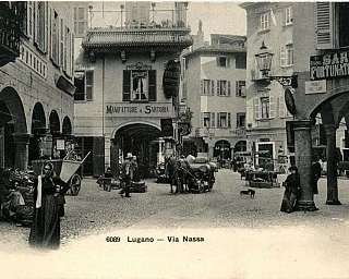 Vue historique de la Via Nassa à Lugano avec des personnes, un chariot tiré par des chevaux et des bâtiments avec des arcades