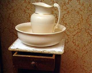 Cruche blanche avec bord doré dans une bassine blanche sur une petite table en bois avec tiroir