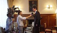 Interview télévisée entre un homme en veste et un journaliste avec un caméraman dans une pièce aménagée