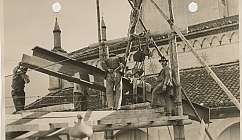 Quatre ouvriers travaillent sur un échafaudage en bois avec des poutres métalliques devant un bâtiment historique