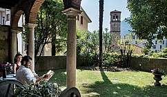 Couple assis sous des arches dans un jardin avec pelouse, plantes et clocher en arrière-plan