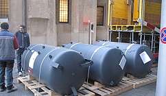 Trois réservoirs cylindriques gris sur une palette en bois avec deux hommes à proximité dans un environnement urbain