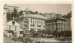 Photo en noir et blanc de bâtiments historiques et de végétation le long de la rive d'un lac avec des personnes et des voitures anciennes