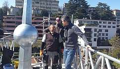 Trois hommes souriants sur un échafaudage métallique avec des bâtiments et des arbres en arrière-plan