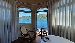 Intérieur d'une chambre avec fauteuil et table, vue sur le lac et les montagnes depuis la fenêtre et la porte ouverte