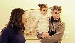 Homme tenant une fillette dans ses bras tandis qu'une femme les regarde dans une pièce avec un tableau