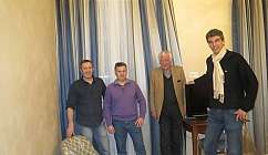 Quatre hommes debout dans un salon avec des rideaux bleus et des meubles en bois