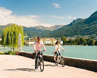 Zwei Personen auf dem Fahrrad entlang eines Sees mit Bergen im Hintergrund.