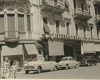 Scene of a street with vintage cars and people walking.