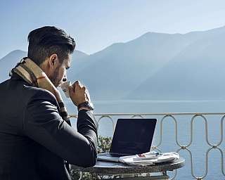Man in a jacket drinking coffee in front of a laptop with a mountain view.