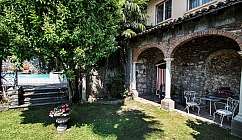 Giardino con prato, albero, piscina, portico con archi in mattoni e sedie bianche