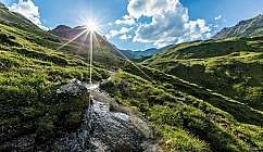 Sentier avec ruisseau entre collines verdoyantes et soleil éclatant derrière la montagne