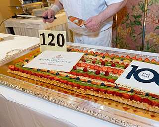 Torta rettangolare decorata con frutta su un vassoio, mentre una persona la rifinisce